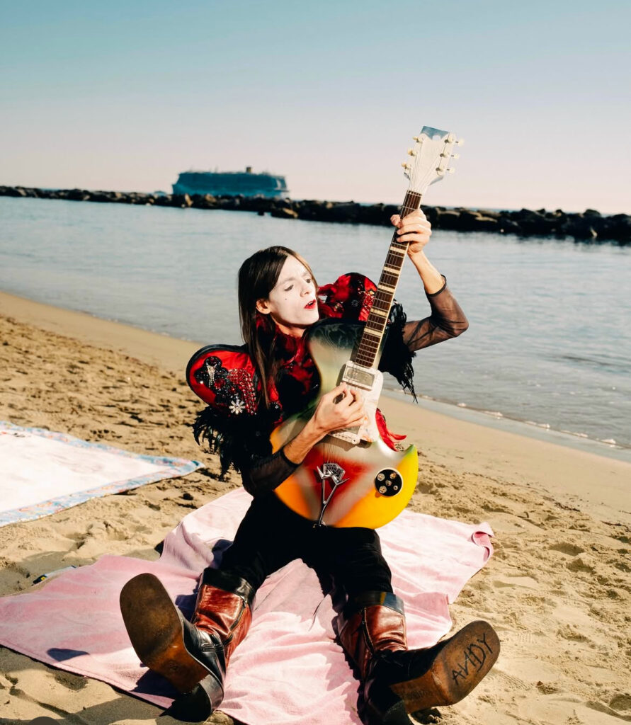 Lucio Corsi seduto sulla spiaggia suona la chitarra con una maglia con spalline vaporose